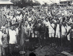 A couple hundred men are all facing the camera, smiling and cheering. Many have their hands raised. The men are wearing uniforms, t-shirts, and shorts. Huts and trees can be seen in the background.