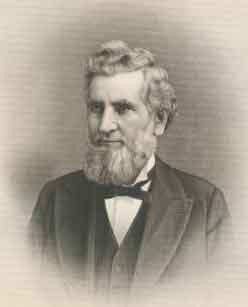 An older man, facing left, with curly, white hair and a beard. He is wearing a white shirt, black tie, and black jacket