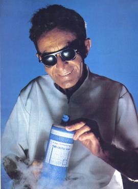 A picture of a blind man wearing dark glasses and a white lab coat grinning while holding a bottle of soap while lit from below. Steam, presumably dry ice, billows from below, giving the photograph a science-fiction feel.