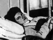 A white woman with dark hair is lying in a hospital bed; she is looking at the camera