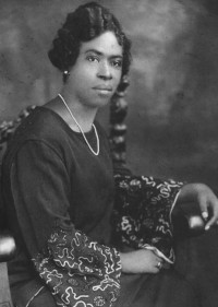 studio black and white photograph of woman posed on chair wearing dress, pearl necklace, and earrings