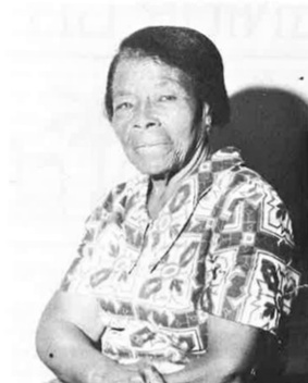 1/2 portrait photograph of a black woman in a patterned, short-sleeved dress whose arms are crossed in her lap.