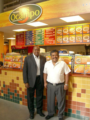 Two men, a taller one on the left in an open suit jacket and the shorter one on the right in a white button-down shirt, smile as they stand in front of an indoor taquería.