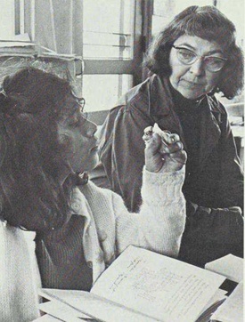 A photograph of a student examining something held in her left hand sitting at a desk with an open book on it and flanked on the left by her teacher who is wearing glasses.