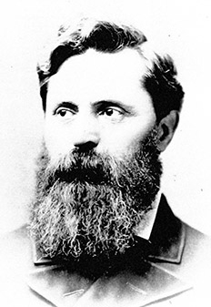 A greyscale portrait photograph of a white man with a large beard in a coat; he is facing and looking to the camera's left.