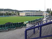 Area of mown grass with rugby posts and a white fronted pavilion building. In the foreground are terraces and seating with hills in the distance.