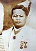 Middle aged Filipino American male wearing a white suite with a neck order medal, and two other medals on the lapel.