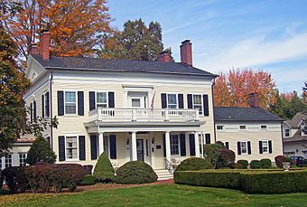 A pale yellow house with white trim, a black roof and shutters, for brick chimneys and a front porch with square pillars and a balcony on top. There is a lawn with manicured shrubbery in front, and behind it there are trees, most of which are in autumn color