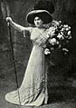  Woman standing in a dramatic pose, right arm raised, left arm holding a large bouquet. She is wearing a long formal gown and a wide-brimmed hat.