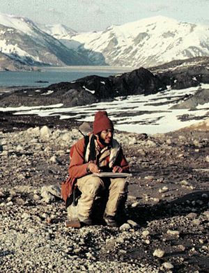 Man wearing a coat and hat and holding a pad of paper sits on a rock, with a lake and several mountains visible in the background