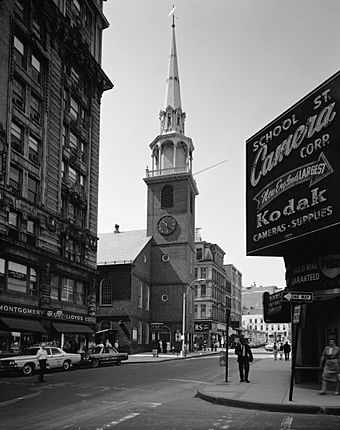 On a city street, an old brick church with a tall steeple is flanked by modern buildings.