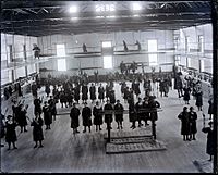 Group of Young Women Students in School Uniform at Gym Class; Some with Indian Clubs; Some on Parallel Bars, Others on Catwalk Above Gym Floor 1880
