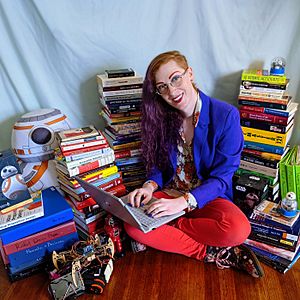 A woman sits on the floor with a laptop surrounded by books and robots