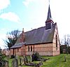 A red brick church with a steeply sloping slate roof seen from the northwest; at the right is a bellcote with a spirelet, and the vestry with its chimney protrudes from the body of the church