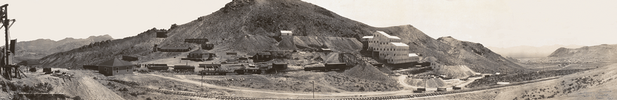 A large mill, five or six stories high and built on the side of a mountain, is surrounded by mine tailings, rail cars, piles of lumber, and a variety of smaller buildings. A rail line leads from the mining operation toward a city nestled between hills in the distance.