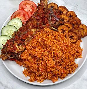Jollof rice with fried fish and plantains garnished with cucumber and tomatoes
