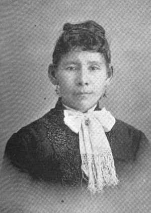 A Native American woman in a formal portrait. Her hair is arranged up and back, away from her face and shoulders, ears exposed; she is wearing a dark jacket with a light-colored fringed fabric bow at the neck.