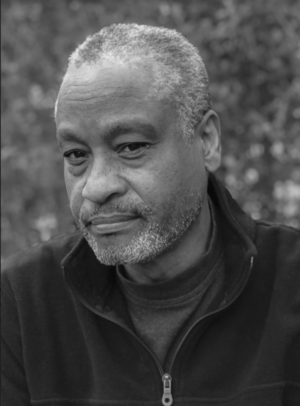 The black and white photograph depicts Ron Tarver, a Black man, looking right at the camera. He has closely cropped salt and pepper hair and stubble, and is wearing a black zip-up jacket.