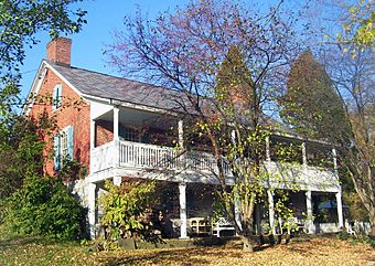 A brick house with full-width wooden porch and balcony seen from the left. A tree that has lost most of its leaves is in front, with other shrubs and trees at the side and rear.