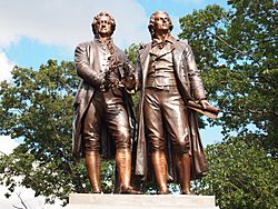 Photograph of a statue of Goethe and Schiller standing side by side, each looking forward. There are trees and blue sky visible behind the statue. The two figures are of nearly the same height. Goethe appears middle-aged; Schiller is noticeably younger. They are dressed in nineteenth century clothing. Goethe is wearing a knee-length formal coat, and Schiller is wearing a calf-length coat. Both men wear breeches. Goethe's left hand rests lightly on Schiller's shoulder; his right hand holds a laurel wreath near his waist. Schiller's right hand is nearly touching the wreath, which may suggest that Goethe is passing the wreath to Schiller. Schiller's left hand extends loosely below his waist, and grasps a rolled sheet of paper.