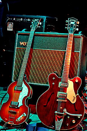 to elektriske guitarer, en lysebrun violinformet bas og en mørkere brun guitar, hviler mod en Voksforstærker.