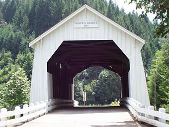 A narrow road leads through a white covered bridge toward a forested hill. The bridge is about 25 feet wide, 25 feet tall to the peak of its roof, and 100 feet long. White guardrails line both sides of the road and the floor of the bridge. A sign above the bridge portal reads, "Hayden Bridge 1918".