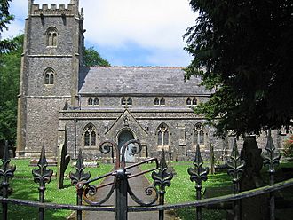 Gray stone building with square tower at left hand end. Foreground includes grass area with gravestones, taken over the top of metal railings.
