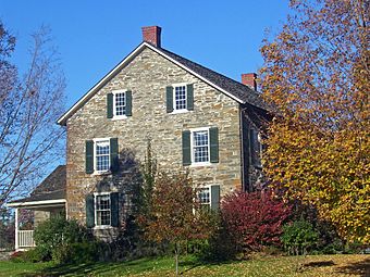 Side view of a stone house with pointed roof, two chimneys and a small wing on the left. On the right is a tree whose leaves are changing with autumn.