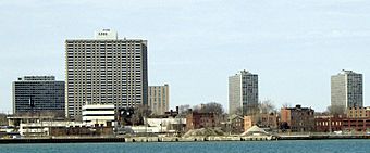 Several modernist gray high-rises with smaller, brown brick buildings beneath the bare trees among them, seen from across a body of water