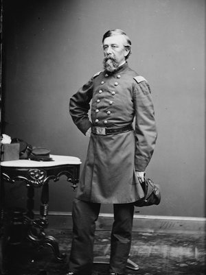 Bearded, older man stands in full military uniform, holding a kepi.