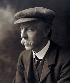 Black and white portrait photograph of Sir Edward Albert Sharpey-Schafer, looking to the left, wearing a flat cap and suit. He was a large moustache.