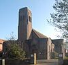 A small brown brick structure unrelieved by dressings or other ornamentation, and partly obscured by shadow, trees and a noticeboard. In the centre is a relatively tall, narrow tower with two arched openings near the top.