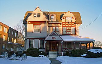 An intricate three-story beige wooden house with red trim, with snow on the porch roof and the front lawn, with a setting sun lighting the upper stories from the right. It has windows of different shapes and sizes. A front porch is on the right half of the first story, with a triangular projection on the left with a prominent "36" in the center. Another triangular section projects from the left hand side of the roof, with a smaller projection with a segmented, more curvilinear roofline to its right. A brick building is visible on the left; in the left front of the yard is an old horse-drawn carriage.