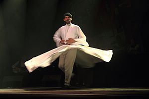 Mercan Dede, wearing a white outfit and dark green turban, whirling onstage