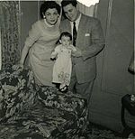 A woman and man, both in their thirties and both dressed in 'Sunday best', hold a similarly dressed very young girl standing on the arm of a floral-print sofa.