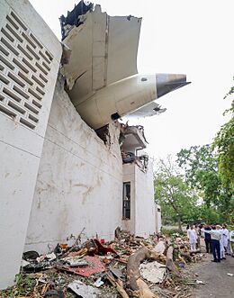 The tail section and one landing gear of an aircraft visible protruding from the upper section of a two-storey building. At the ground is a pile of rubble containing concrete, steel, and tree branches. Nearby, a man with his back to the camera points at the aircraft while facing Prime Minister Narendra Modi.