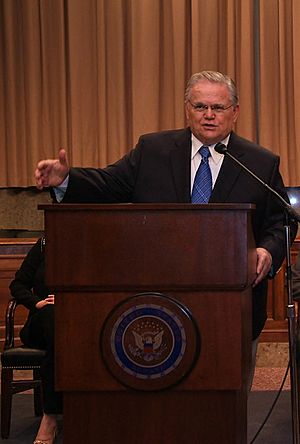 Clean-shaven man in his 60s, with gray hair, wearing glasses, dressed in a dark suit and blue tie, speaking from behind a dark, varnished wooden lectern, with his right arm outstretched. The front of the lectern is emblazoned with the Great Seal of the United States.