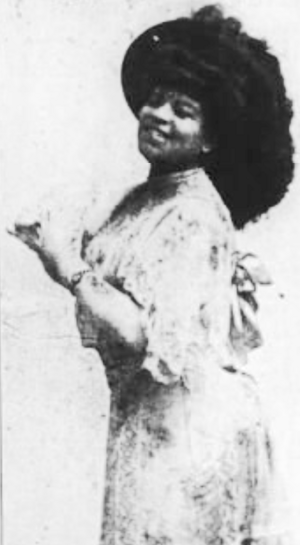 An African-American woman, standing and smiling with hands at her chest; she is wearing a light-colored gown and a large dark hat
