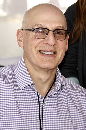Black and white photograph of Gordon Korman, author of young adult fiction, speaking at the National Book Festival in September 2011. The photograph depicts Korman in profile view, facing left and speaking into a microphone. His right hand is raised to approximately shoulder height, palm facing the audience, with fingers slightly closed as if grasping an invisible ball.