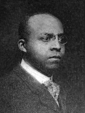 An African-American man with close-cut hair wearing glasses, a black jacket, light shirt, and patterned tie