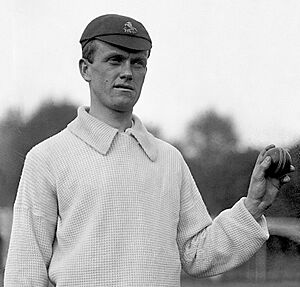 A head and shoulders black and white photograph of a young man wearing cricket whites and a cap and holding a cricket ball in his left hand