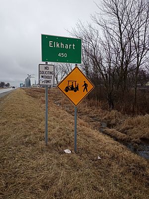 Large green sign in foreground reading "Elkhart, 450", mounted on two metal posts, with a smaller white sign reading "No soliciting without permit" and a yellow diamond-shaped sign with a drawing of a forklift and a person walking; grain elevator in background