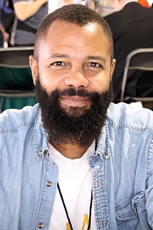 A headshot of Javaka Steptoe smiling at the camera. He has short hair and a beard, and wears a blue jean shirt over a white t-shirt.