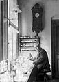 An old, bespectacled man wearing a suit and sitting at a bench by a large window. The bench is covered with small bottles and test tubes. On the wall behind him is a large old-fashioned clock below frick u  which are four small enclosed shelves on which sit many neatly labelled bottles.