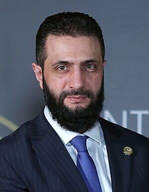 Headshot of al-Sharaa wearing a black suit jacket, a white shirt, and a necktie.