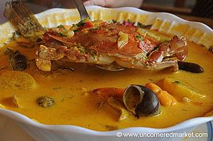 Photo of a bowl of tapado soup, yellow-orange in color, with crab, fish, and other ingredients visible in the bowl
