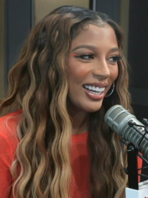 A woman with long, wavy hair is wearing reddish orange and smiling next to a microphone.