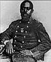 A black man with beard sitting with his right arm propped on an object beside him. He is wearing a double-breasted military jacket with stripes on the sleeve cuffs and a round medal pinned to the center of the chest.
