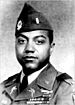 Head and shoulders of a black man with full cheeks in military uniform. Rows of ribbon bars are on his left breast and his lapels and garrison cap are adorned with pins and badges.