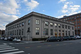 A tall light-colored square two-story stone building with a balustrade and pilasters on the front on an urban street. An American flag flies from a pole on the right. Across the top of the building the words "United States Post Office" is engraved.
